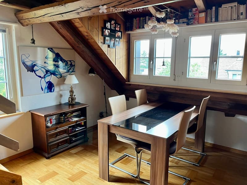 Brightly lit room with wooden floors, large windows, a glass-top desk, chairs, a wooden cabinet, and a lamp on a side table.