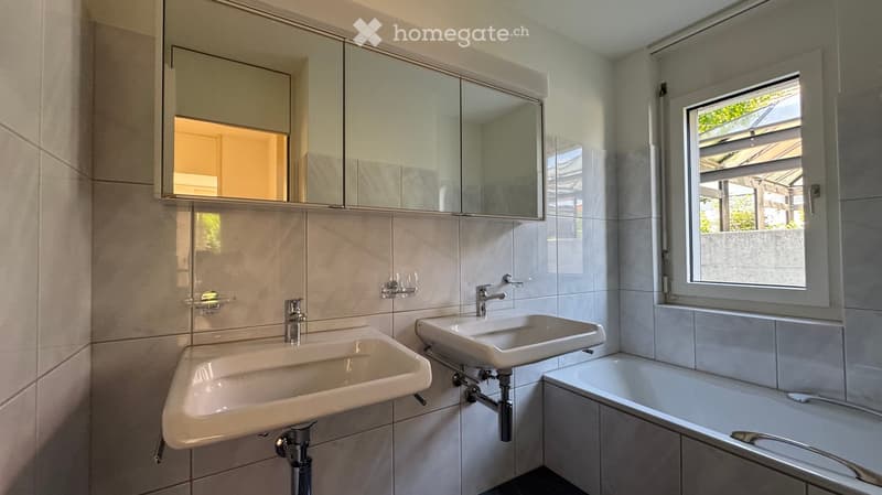 Modern bathroom with two sinks, a bathtub, a large mirrored cabinet, and a window with blinds.