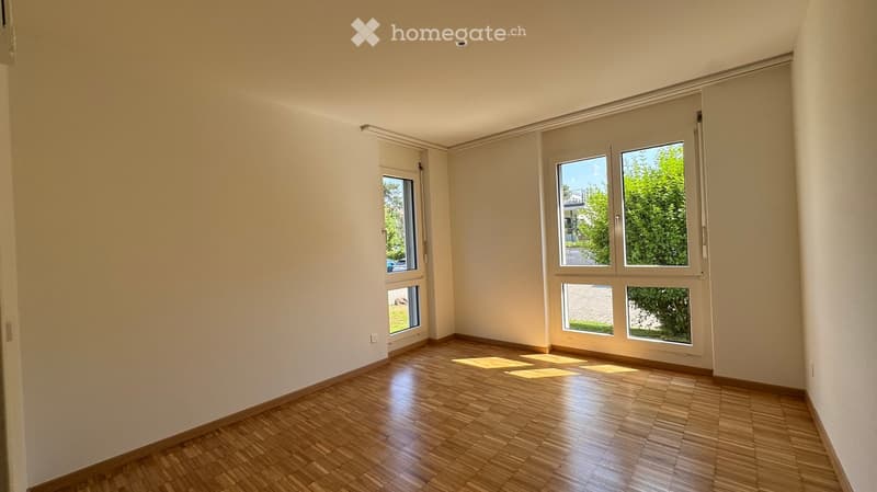 Empty room with wooden flooring and large windows, allowing natural light to fill the space. The walls are painted a light beige color, and the room appears to be well-lit and spacious.