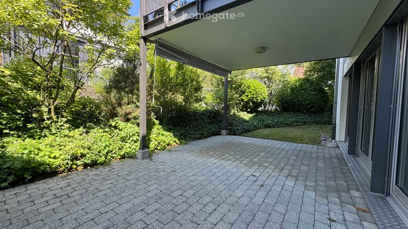 A paved outdoor area with a brick-patterned floor, surrounded by lush greenery and trees, with a modern building structure visible on the right side.