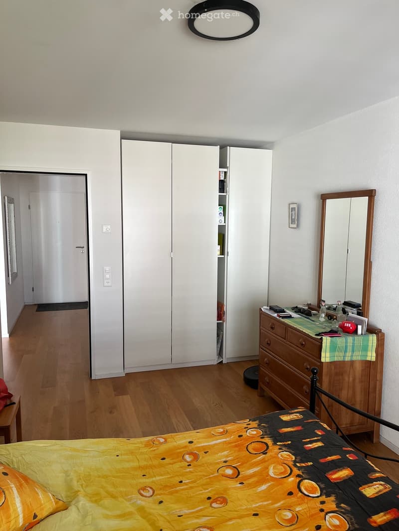 Bedroom with wooden flooring, a bed with a colorful quilt, a large white wardrobe, a dresser with a mirror, and a small table with various items on it.