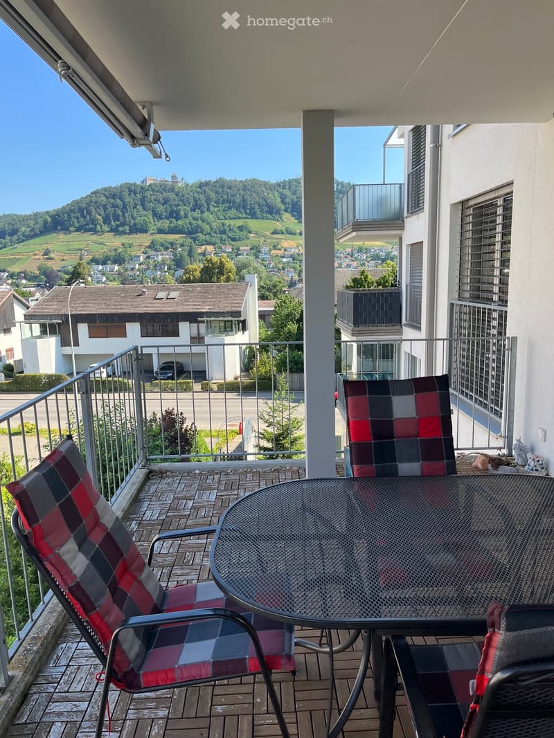 Balcony with a scenic view of a town and hills, featuring a round metal table and two plaid-patterned chairs.