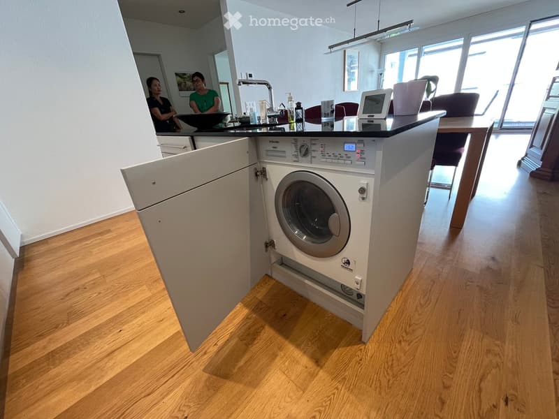 Modern kitchen with light wooden flooring, white cabinets, and a built-in washing machine. A sink with a faucet is visible, along with a table and chairs in the background.