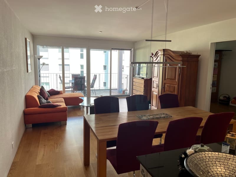 Bright living and dining area with wooden flooring, a sectional sofa, a dining table with chairs, and a balcony with outdoor furniture.