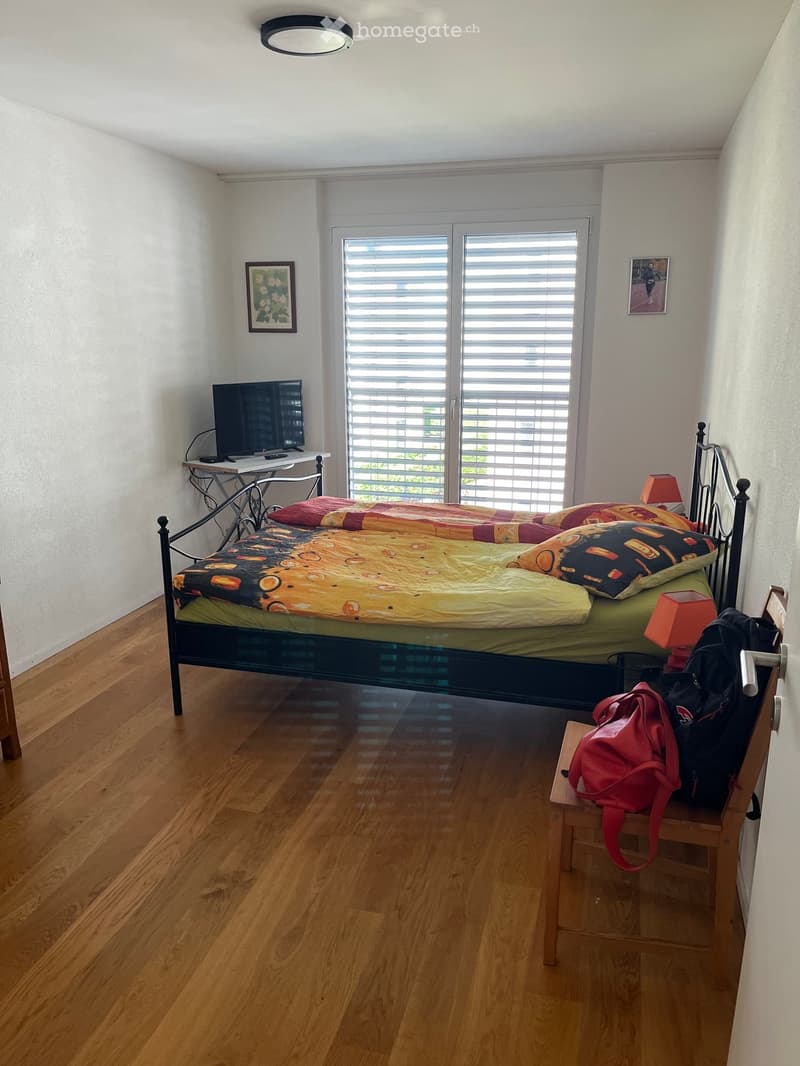 Cozy bedroom with wooden flooring, a bed with colorful bedding, a small table with a screen, and a chair holding a backpack.