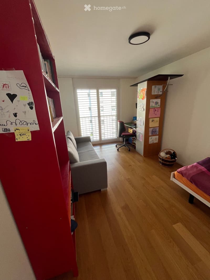 A multi-functional room with a sofa, desk, bed, and shelves. The room features wooden flooring, large windows, and colorful artwork on the walls.