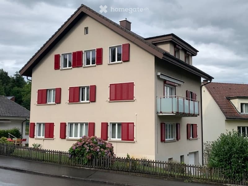 Two-story house with beige walls, red shutters, and a balcony on the upper floor.