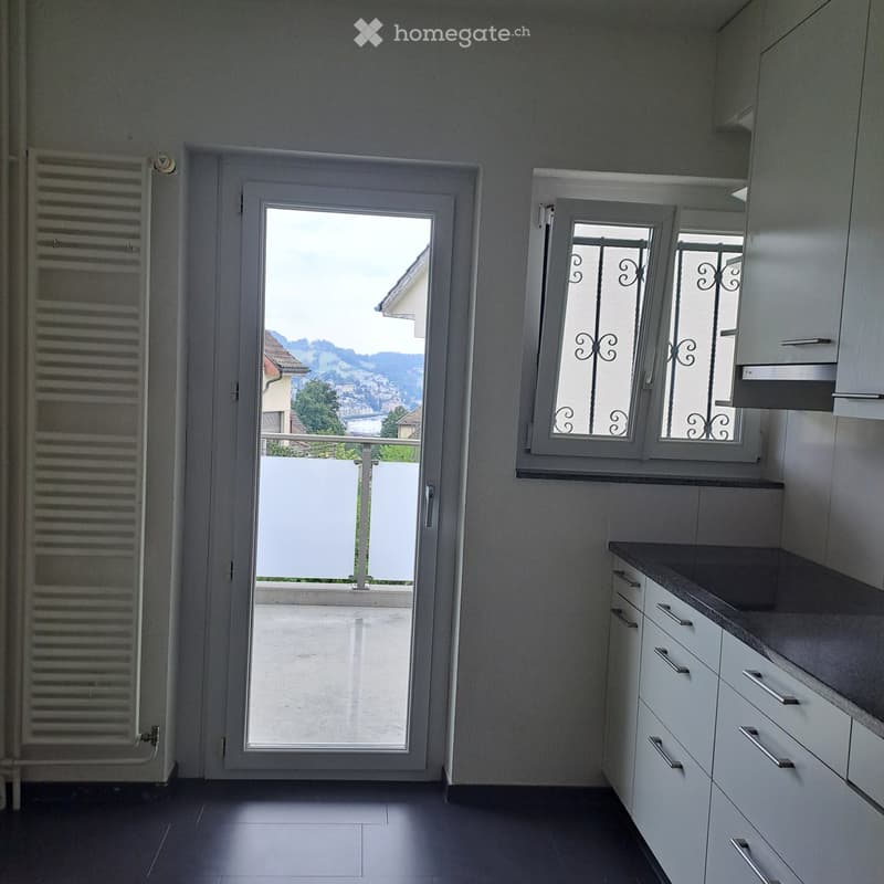 Modern kitchen with white cabinets, dark countertops, and a view of a scenic landscape through a large window and sliding door leading to a balcony.