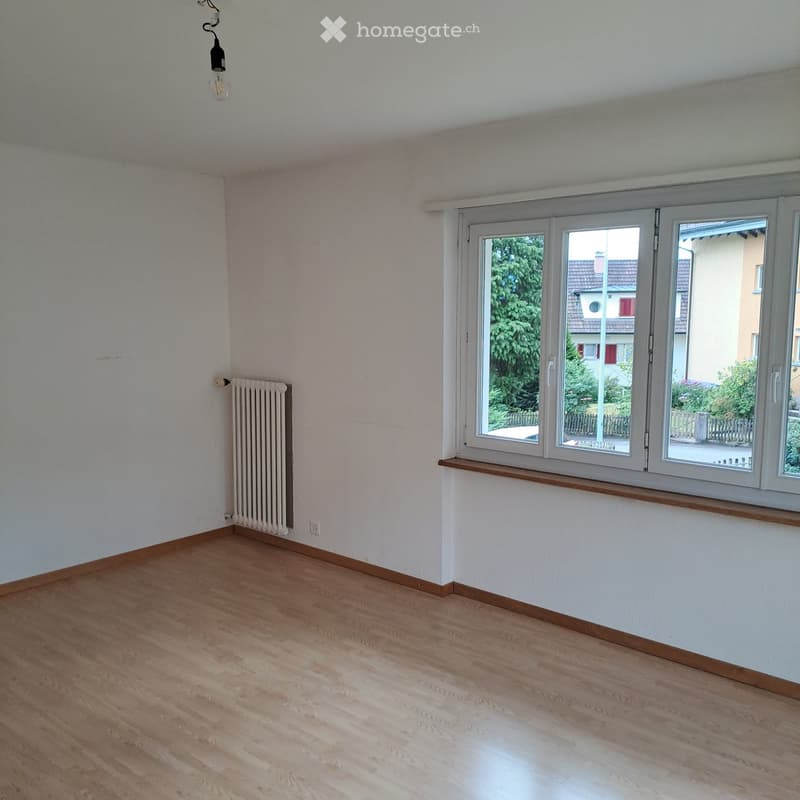 Empty room with light wooden flooring, white walls, large windows with wooden sills, radiator, and a hanging light fixture.