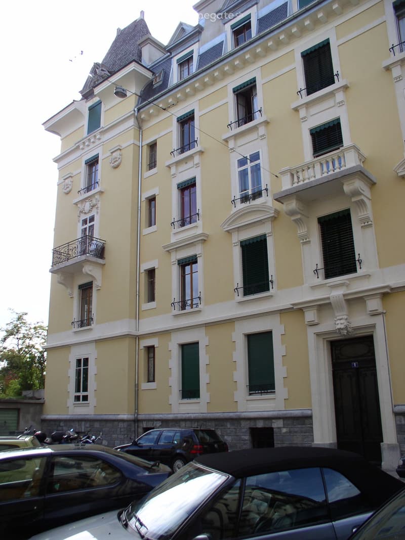 A multi-story yellow building with white trim, ornate balconies, and multiple windows. Several cars are parked in front.