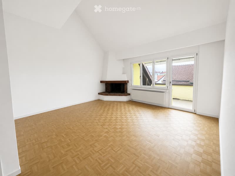 Empty room with white walls, wooden herringbone flooring, fireplace, large windows, and sliding glass door leading to a balcony.