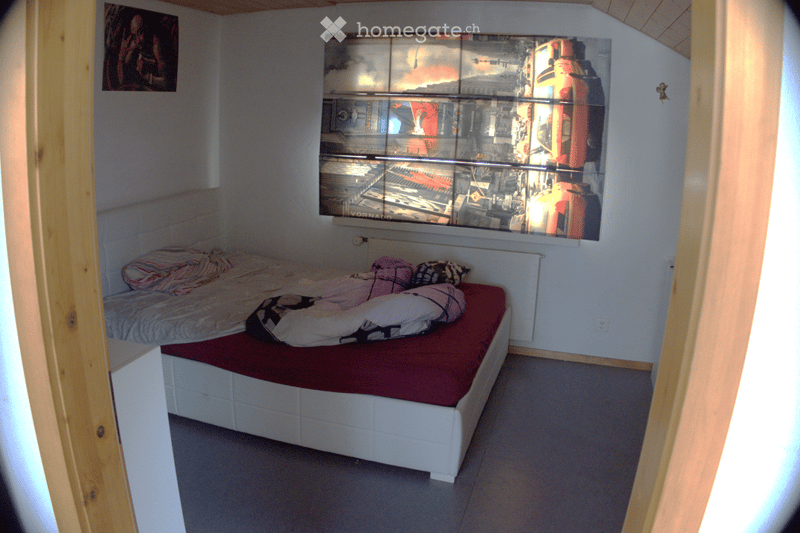 A small bedroom with a white bed frame, red mattress, pillows, and a blanket. A large picture frame hangs on the wall above the bed, and the room has wooden trim and a slanted ceiling.