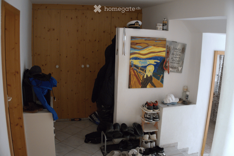 A narrow hallway with a wooden wardrobe, a shoe rack, and various items including jackets, shoes, and a painting on the wall.