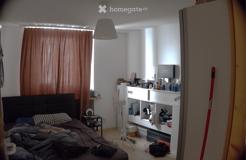 A bedroom with a bed, a fan, a shelf with various items, and a curtain covering a window. The bed is unmade with pillows and blankets.