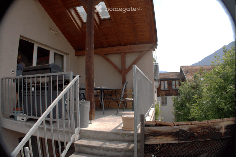 Balcony with wooden roof, metal railing, outdoor furniture including a table and chairs, and a view of mountains in the background.