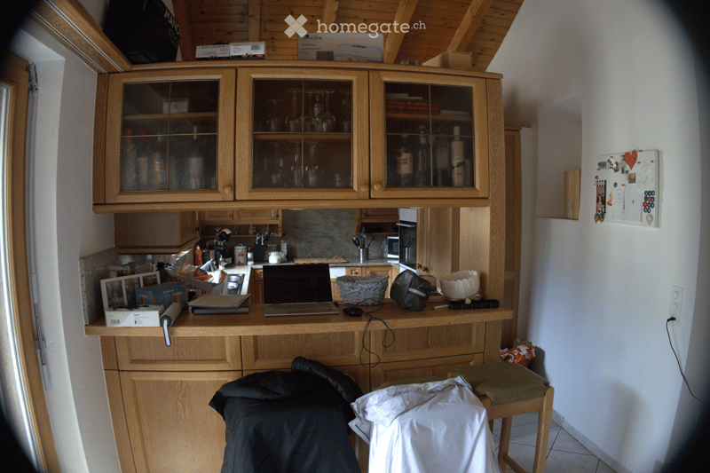 The image shows a small, well-lit room with wooden cabinets and a countertop. On the countertop, there is a laptop, a basket, a bag, and some other small items. There is a stool with a green cushion and a white cloth draped over it. The cabinets have glas