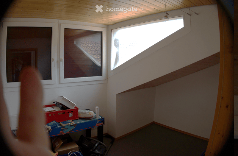 Empty room with wooden ceiling, white walls, and large windows. A small table with storage boxes and a vacuum cleaner is visible, but no furniture or major fixtures are present.
