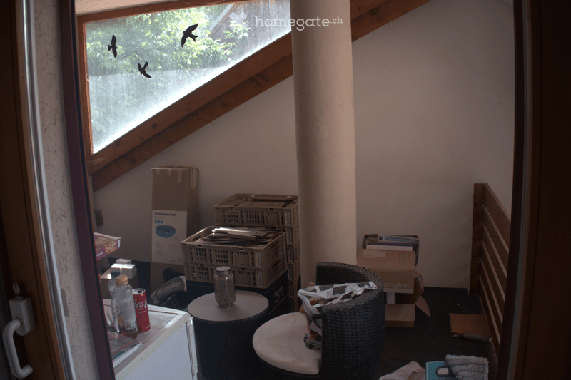 The room features a slanted ceiling with wooden beams, a large window showing greenery outside, and various items including a small round table with a jar, a wicker chair with a cushion, and several stacked cardboard boxes and crates.