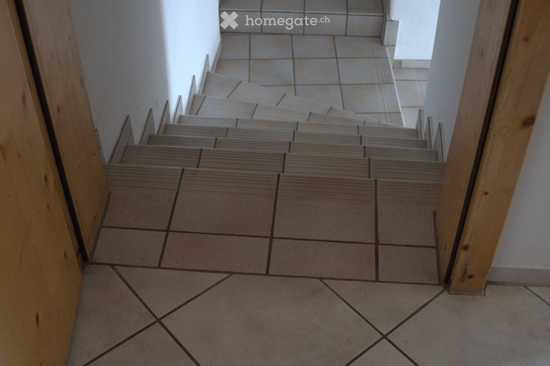 A tiled hallway with a staircase leading upwards, featuring light-colored tiles and wooden railings.
