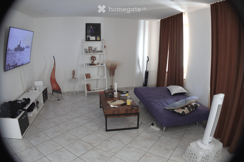 A furnished room with a bed, a table, a chair, a shelf, a fan, a screen, a table lamp, and a curtain.