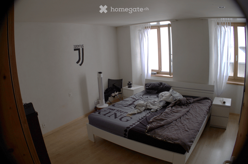 Bedroom with a wooden floor, a bed with a grey duvet, nightstands, a chair, and a fan near the window.