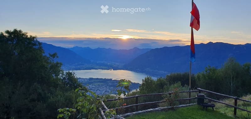 A scenic balcony or terrace overlooking a lake and distant mountains at sunset, with a flagpole and a small wooden fence.