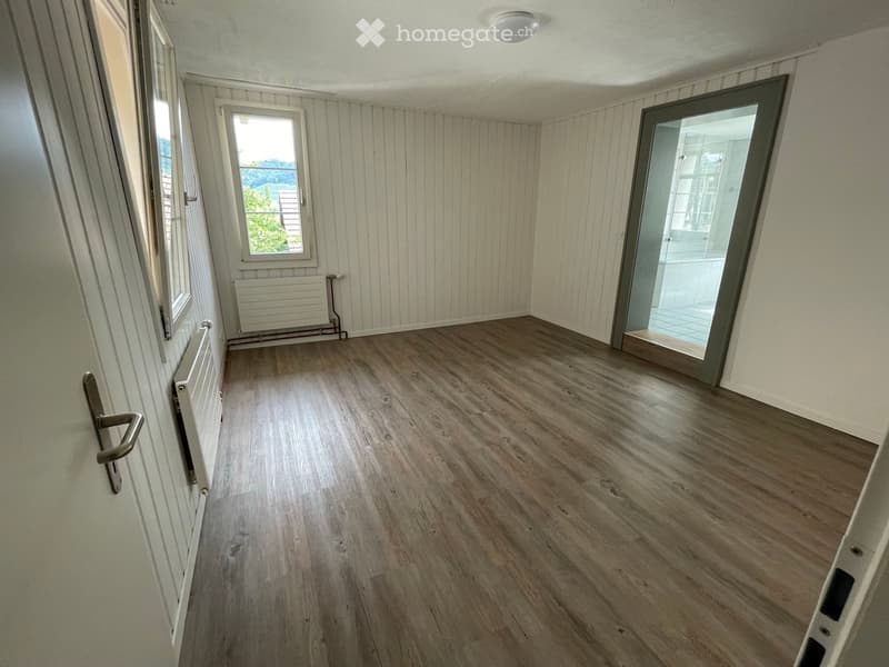 Empty room with wooden flooring, white walls, and a large window with a view of greenery. Features a radiator and a sliding glass door leading to another space.