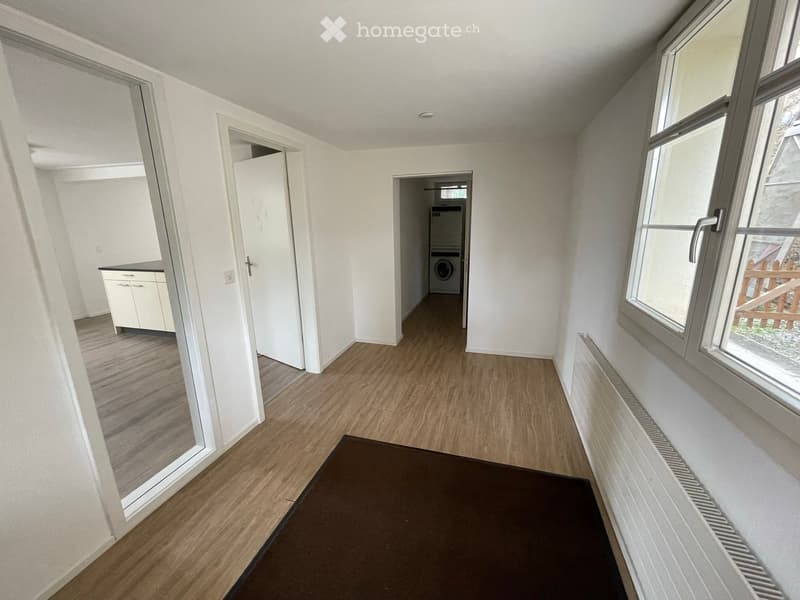 Empty hallway with wooden flooring, white walls, and a radiator. Doors lead to a kitchen area and a laundry room with a washing machine.