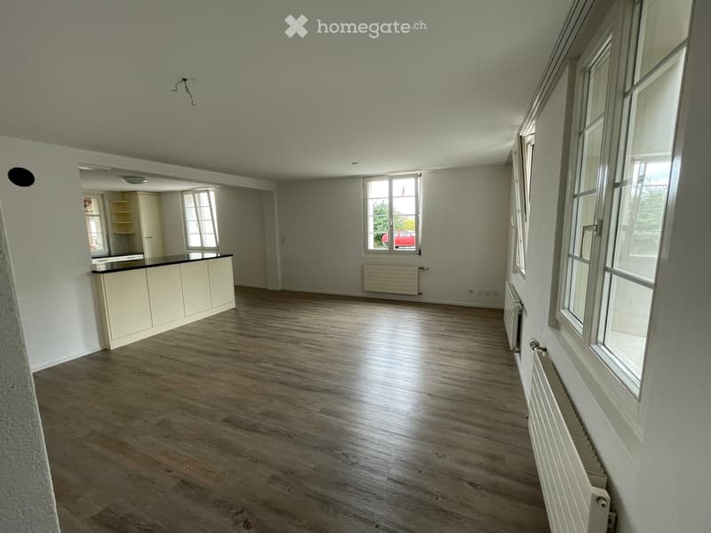 Empty room with wooden flooring, white walls, large windows, and a small kitchen area with cabinets and a countertop.