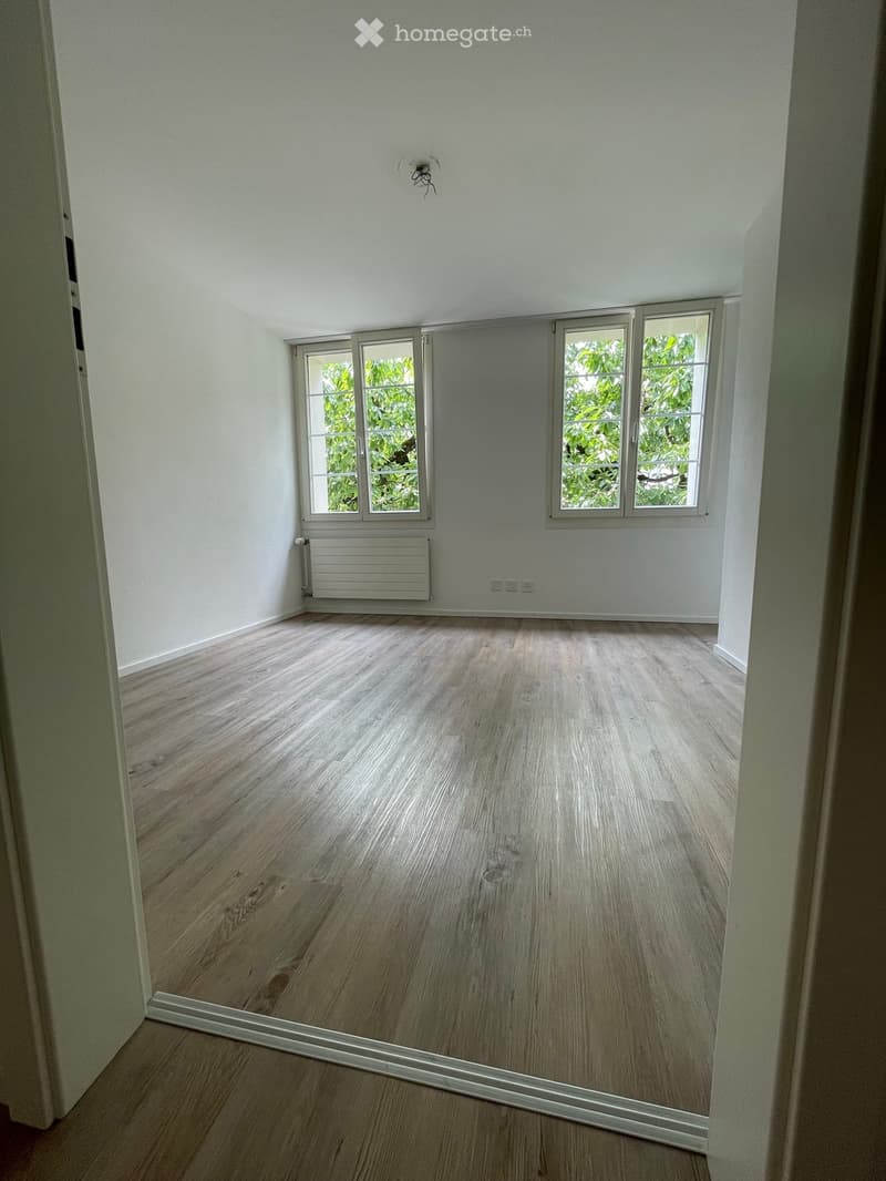 Empty room with light wooden flooring, white walls, and large windows showing greenery outside. No furniture or equipment present.
