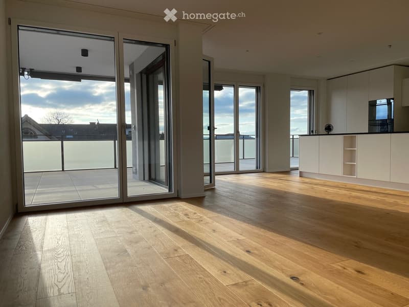 Bright, open-plan space with light wooden flooring, large glass sliding doors leading to a balcony, and a small kitchen area with white cabinets.