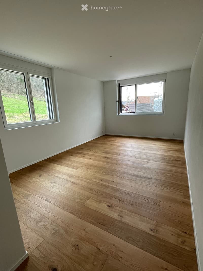 Empty room with light green walls and light wooden flooring, featuring two windows with white frames and a view of a grassy area outside.