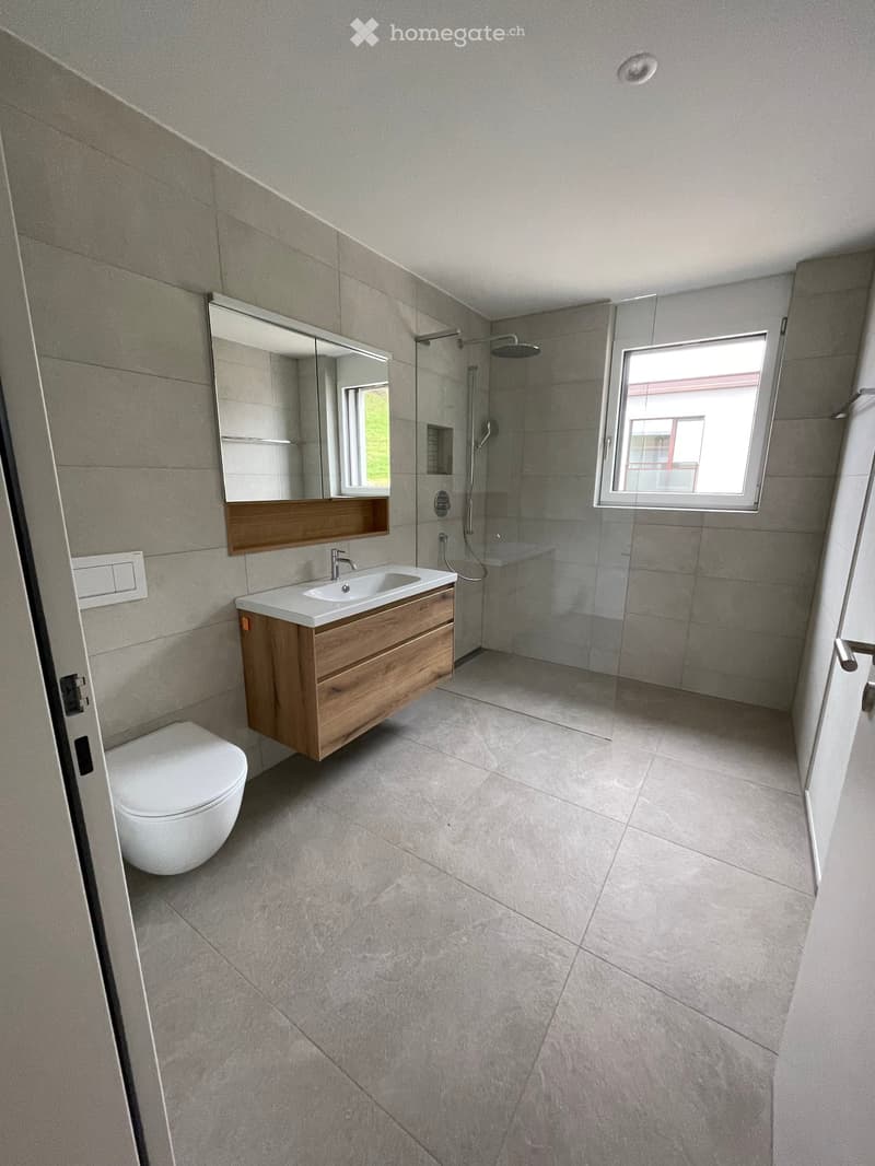 Modern bathroom with a toilet, sink, mirror, and glass-enclosed shower. Features light grey tiles and a wooden vanity.