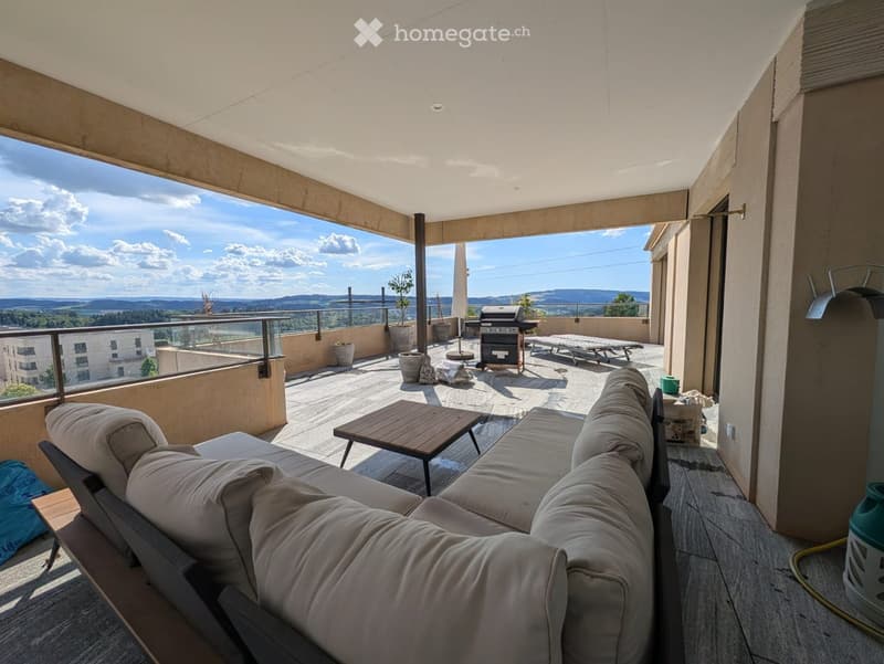 Spacious balcony with a panoramic view, featuring a beige sofa, wooden coffee table, grill, and sun loungers.