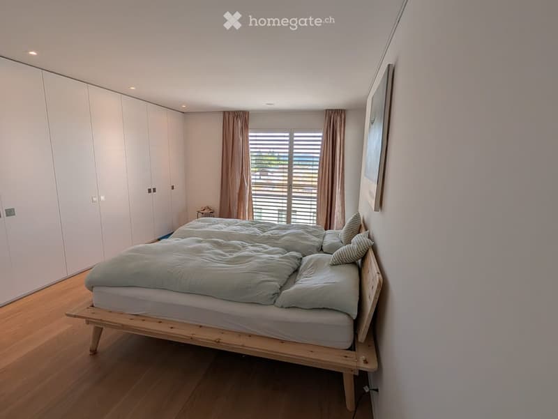 Modern bedroom with a wooden bed, light blue bedding, built-in white wardrobes, and large windows with blinds.