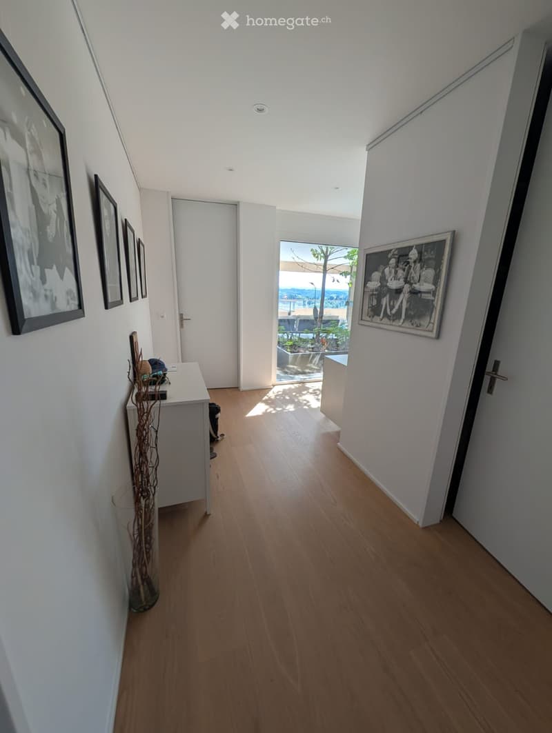 Bright hallway with wooden flooring, white walls, and framed artwork. Features a small console table with decorative items and a view to an outdoor area through a large glass door.
