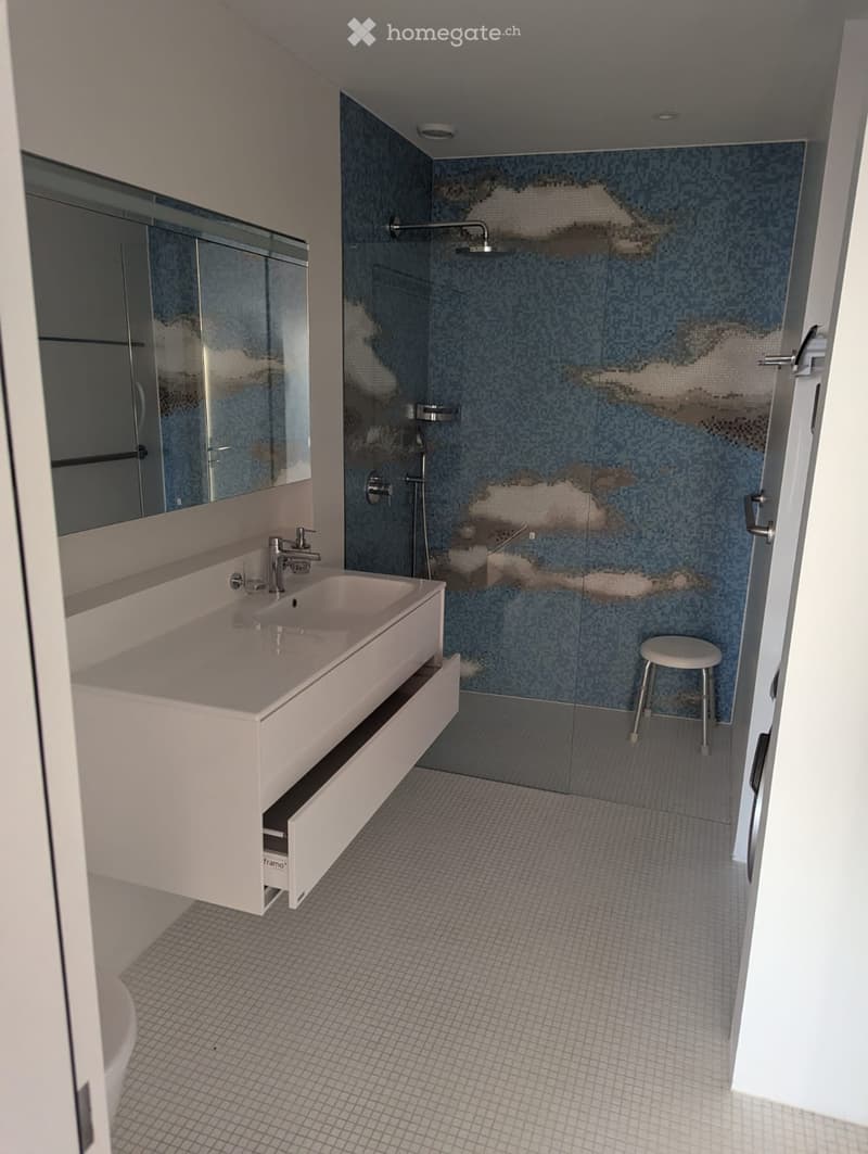 Modern bathroom with a white sink, large mirror, glass-enclosed shower, and a small white stool.