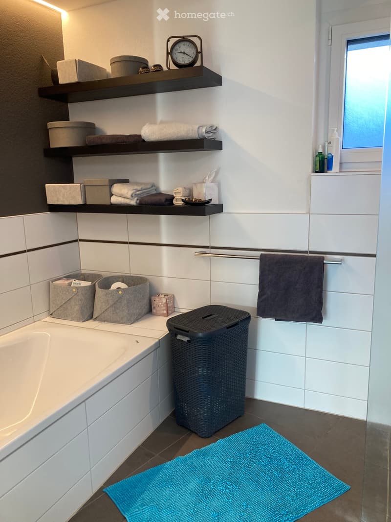 Modern bathroom with white tiled walls, a bathtub, shelves with towels and toiletries, a clock, and a blue bath mat on the floor.