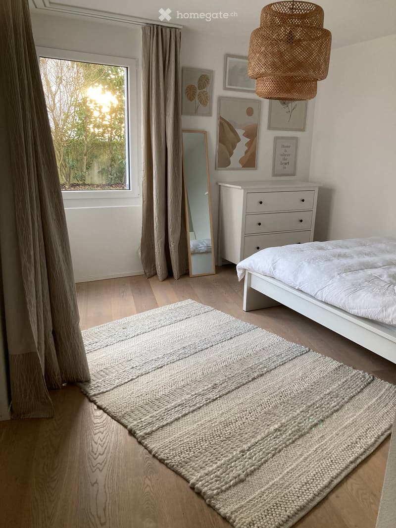 Modern bedroom with a white bed, dresser, mirror, large window with curtains, and a woven rug on the floor.