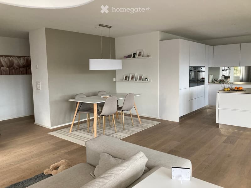Modern open-plan living and dining area with a grey sofa, coffee table, dining table with four chairs, white kitchen cabinets, and a pendant light fixture.