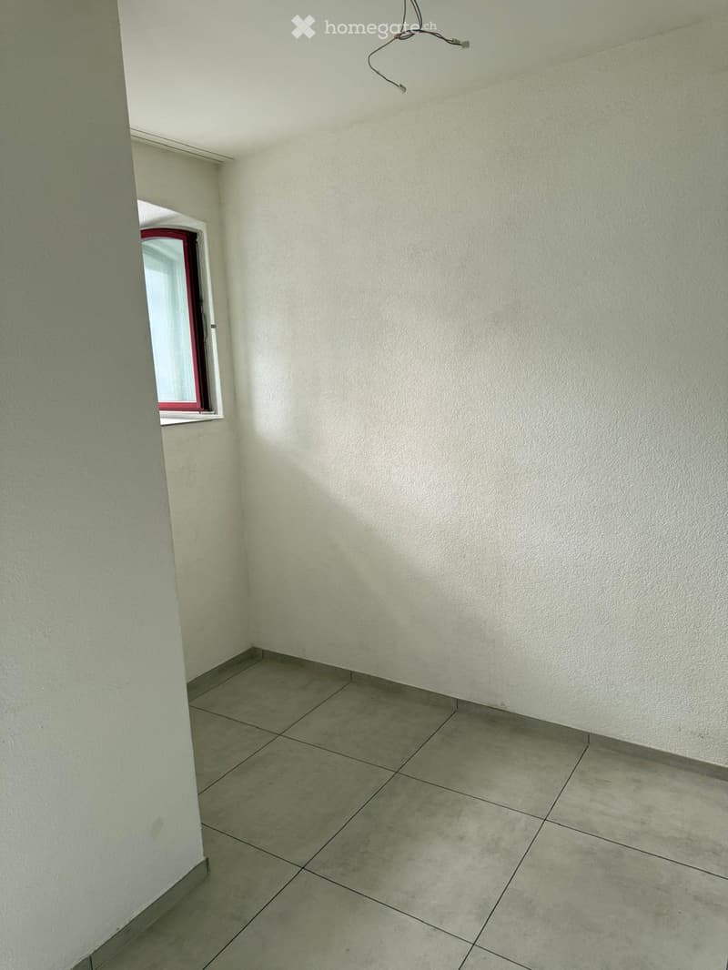 Empty room with light-colored tiled floor and white walls, featuring a small window with red frame and a visible electrical outlet on the ceiling.
