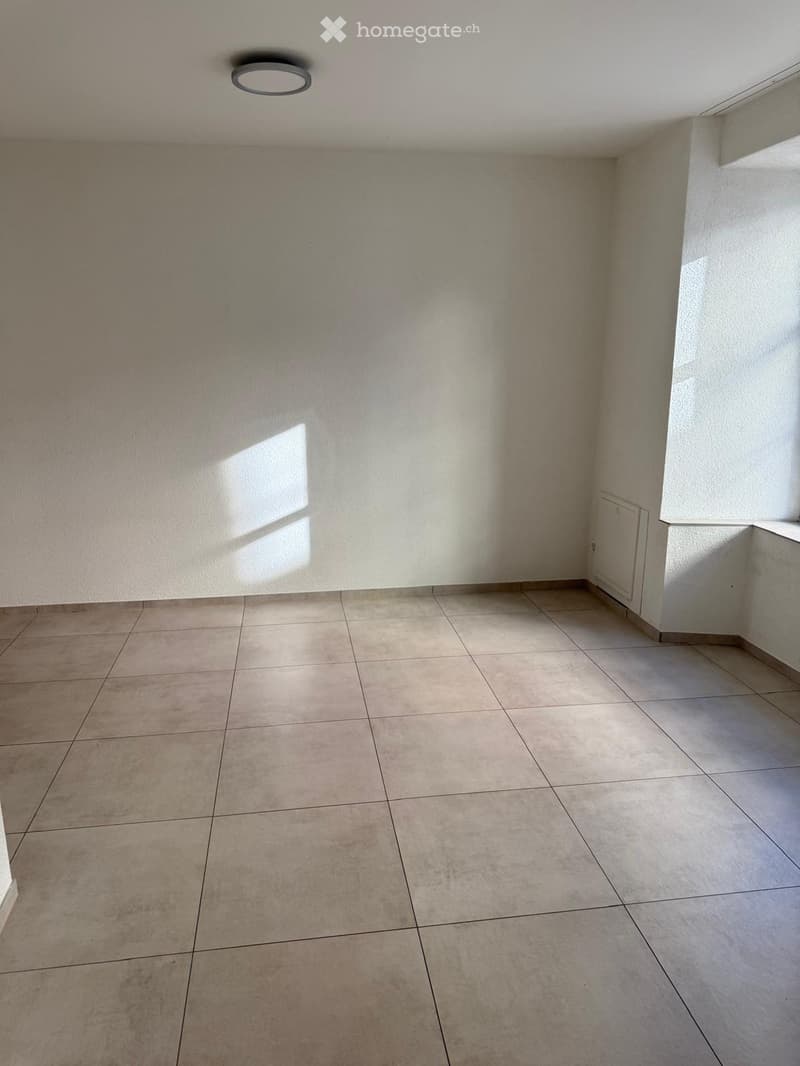 Empty room with tiled floor and white walls, featuring a single ceiling light fixture and a small window allowing natural light.