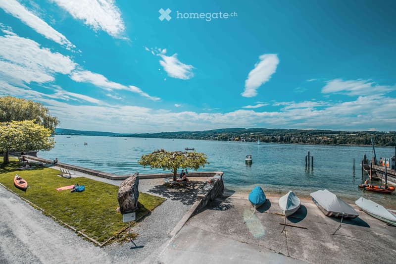 A lakeside park with grassy areas, stone structures, and several boats docked along the shore. People are relaxing on the grass, and the lake stretches out with sailboats and other watercraft visible in the distance.