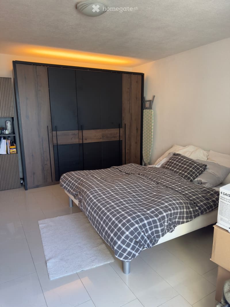 Modern bedroom with a large bed, dark grey wardrobe, nightstand, and a mirror. The room features a white tiled floor and a round ceiling light.