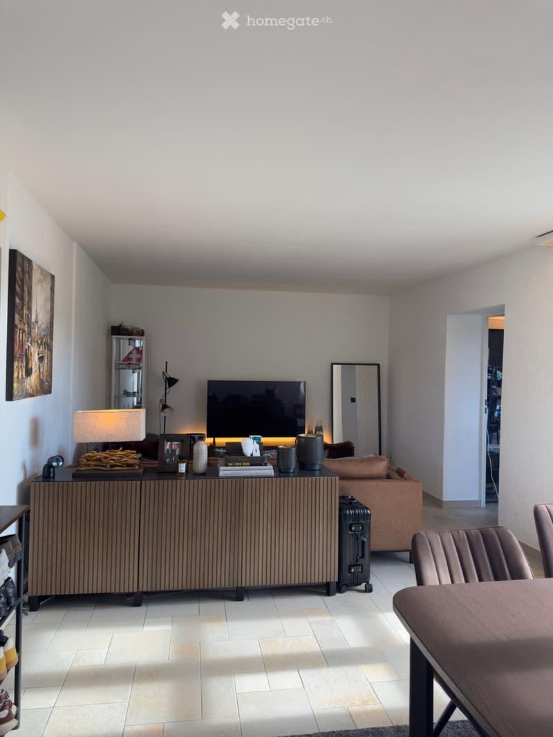 Modern living room with a brown leather sofa, a wooden side table, a lamp, a television, a mirror, and a cabinet. The room features tiled flooring and a slanted ceiling.