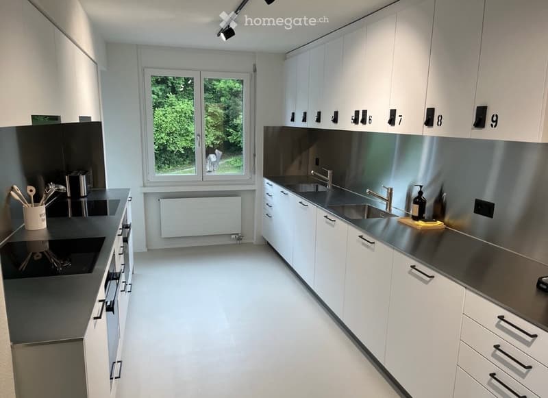 Modern kitchen with white cabinets, stainless steel countertops, and a large window overlooking greenery. Includes a sink, faucet, and wooden utensils in a container.