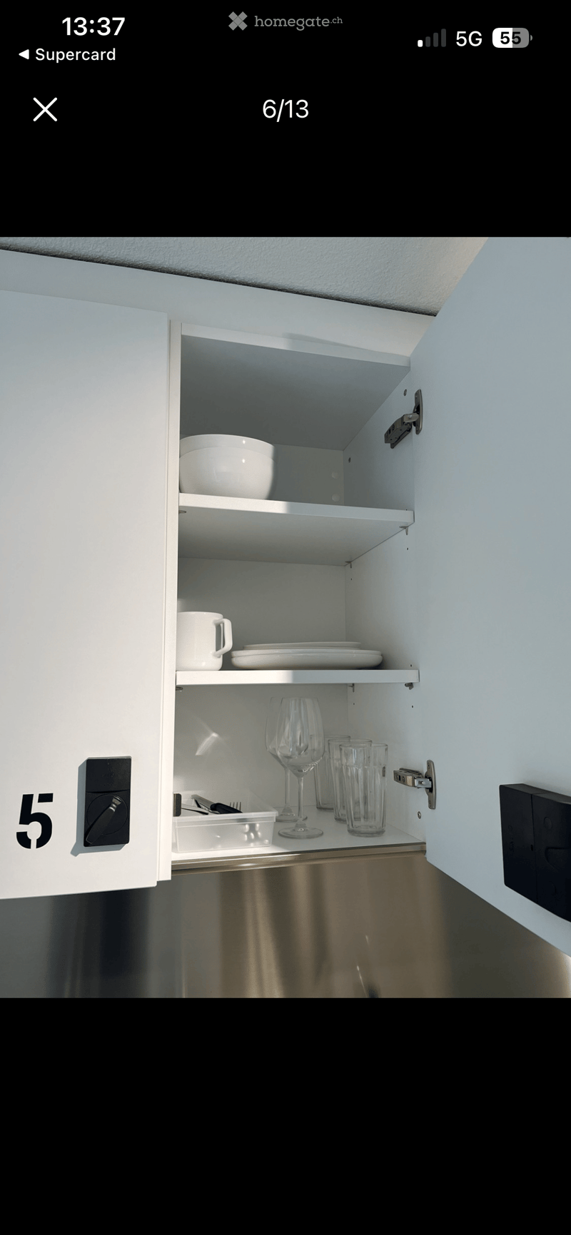 Open kitchen cabinet with white shelves and stainless steel accents, containing a bowl, cup, plates, wine glass, and utensils.