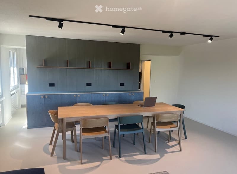 A modern, minimalist kitchen area with a wooden dining table, six chairs, a laptop on the table, and grey cabinets with a sleek design.