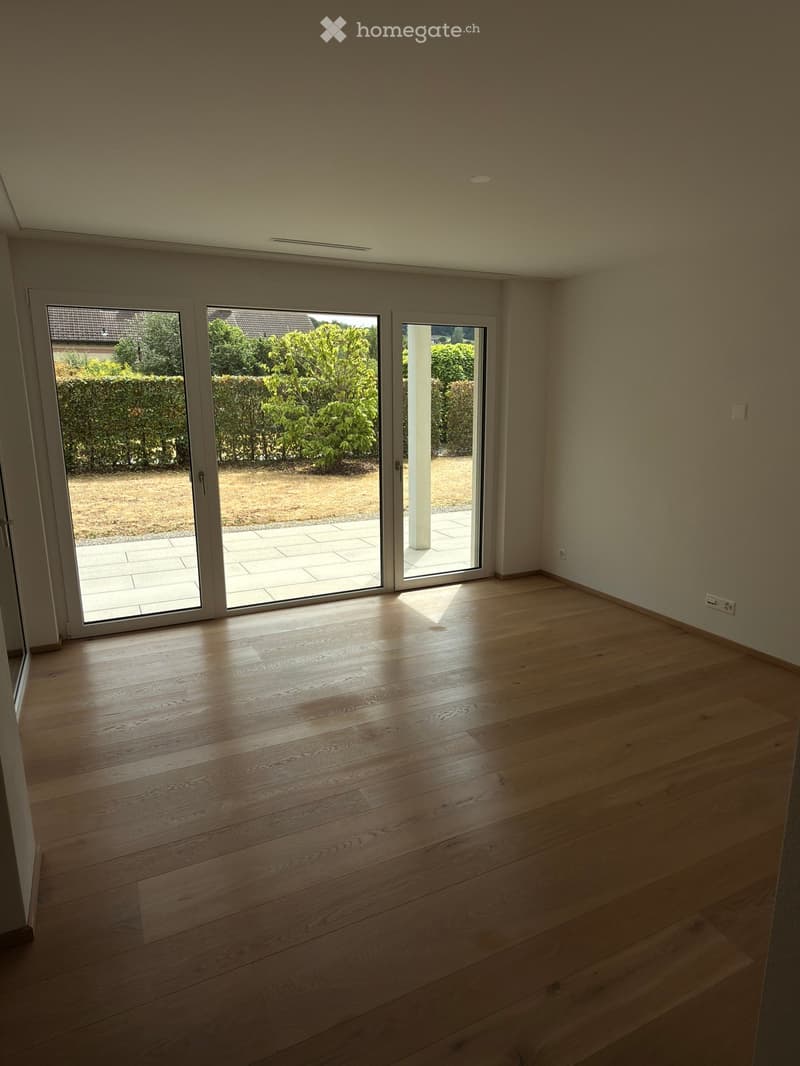 Empty room with wooden flooring and large sliding glass doors leading to a patio area with a view of a garden.