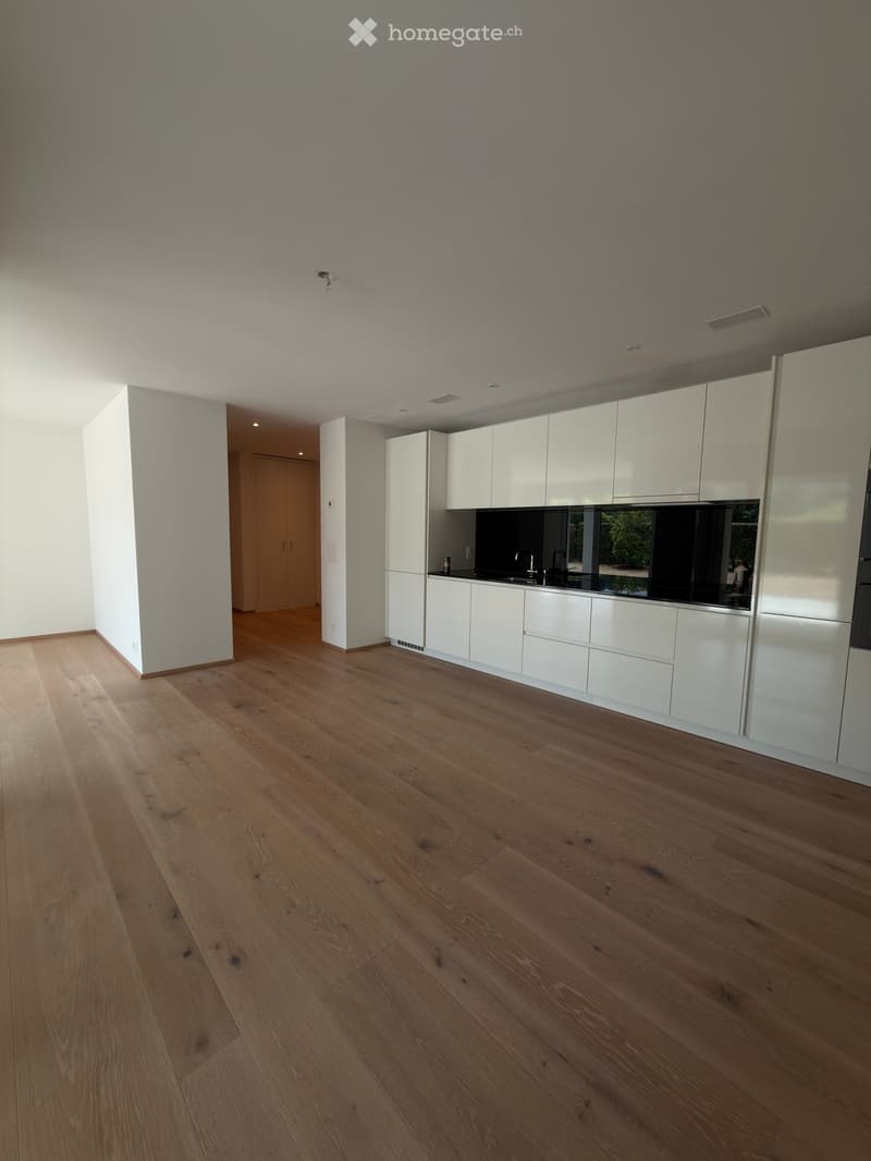 Empty room with light wooden flooring, white walls, and a white kitchen area with cabinets and a countertop visible in the background.